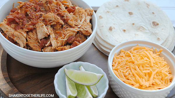 Slow Cooker Mexican Shredded Chicken in white bowl with shredded cheese, lime wedges, and tortillas