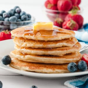 pancakes in stack on white plate with butter and syrup and strawberries and blueberries
