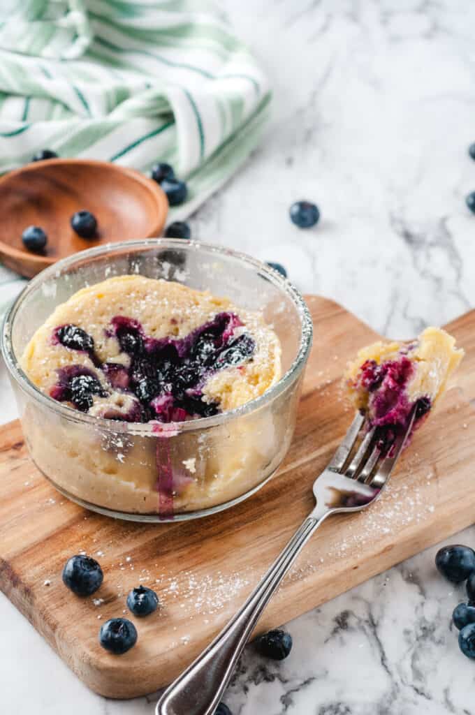 blueberry mug cake in clear glass jar on cutting board with fork holding one bite