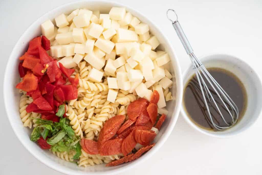 cold italian pasta salad ingredients from overhead in a white bowl with small bowl of dressing with whisk nearby