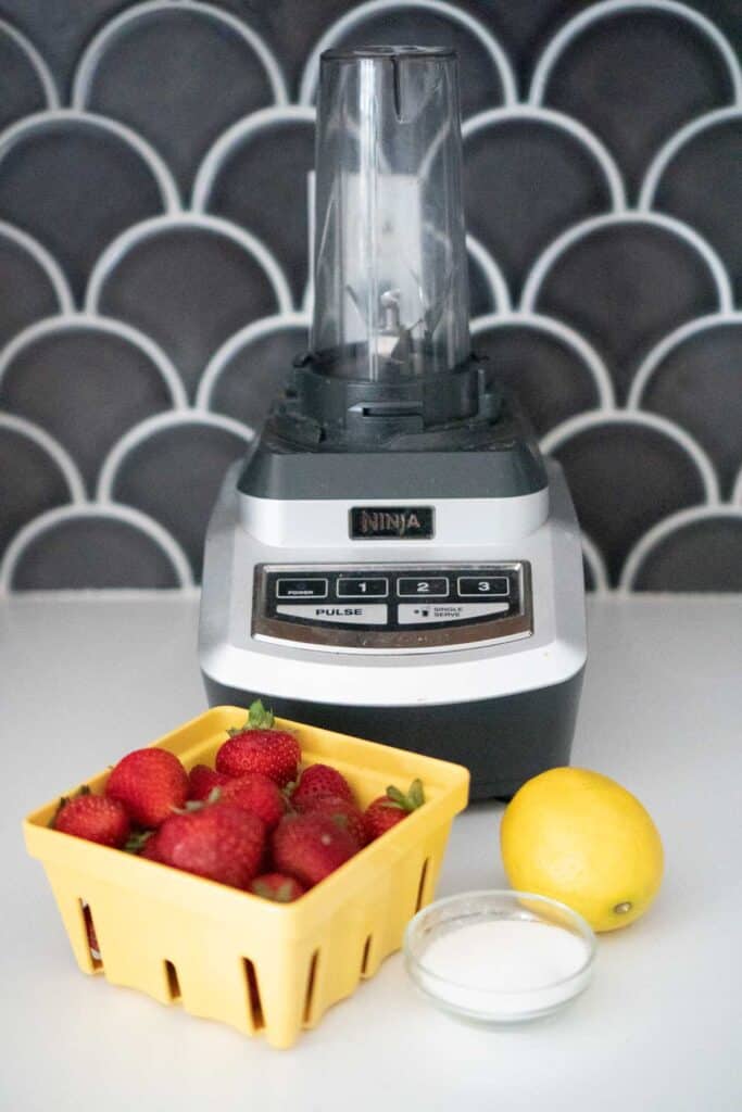 blender with carton of strawberries, lemon and bowl of sugar