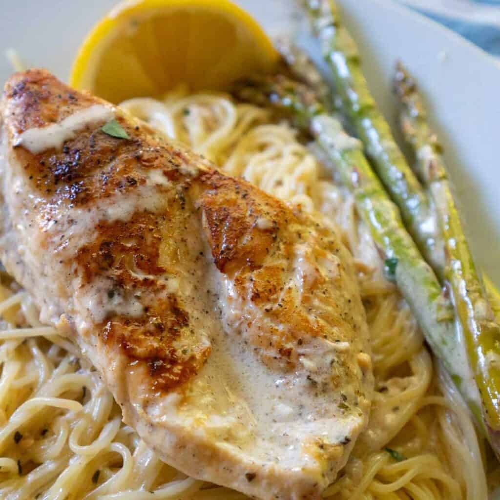 white plate with pasta topped with chicken breast, asparagus and cream sauce and lemon garnish