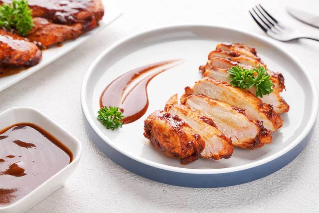 sliced barbecue chicken breast on white plate with extra sauce garnish