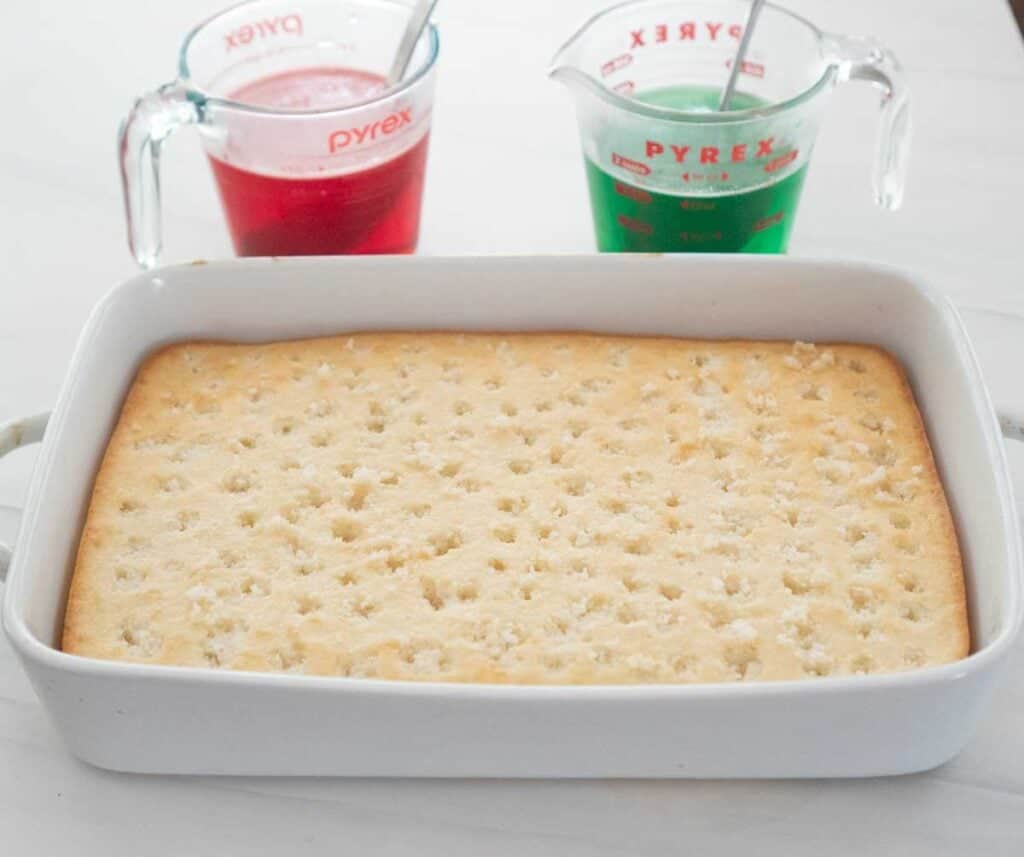 cake with holes poked in the top and two glass measuring cups with red and green dissolved gelatin