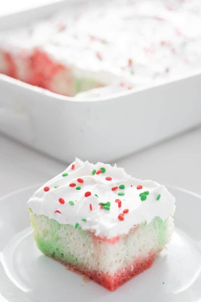 piece of Christmas jello poke cake on white plate in front of the remaining cake
