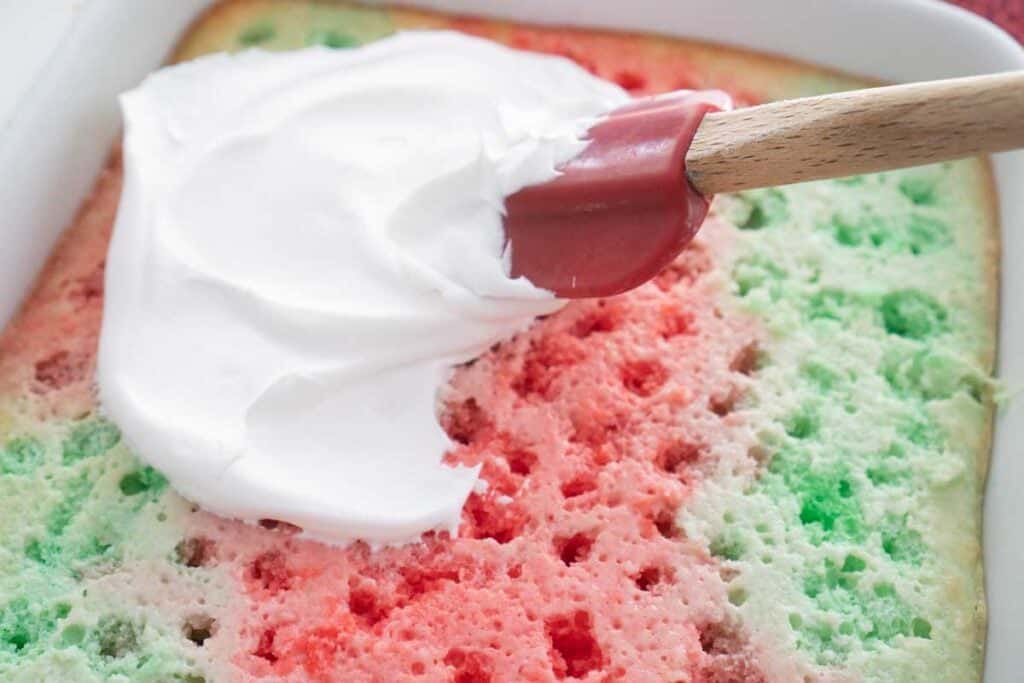 rubber spatula spreading whipped cream over Christmas poke cake