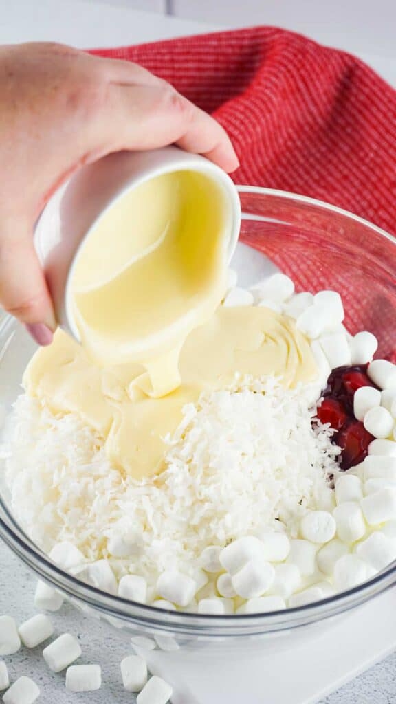 pouring condensed milk into cherry fluff ingredients in glass bowl