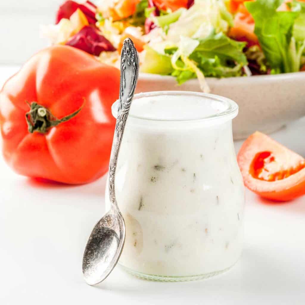 ranch dressing in jar with silver spoon and salad and tomato in background