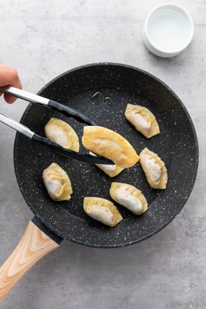 tongs holding pan fried pork dumpling over pan of dumplings